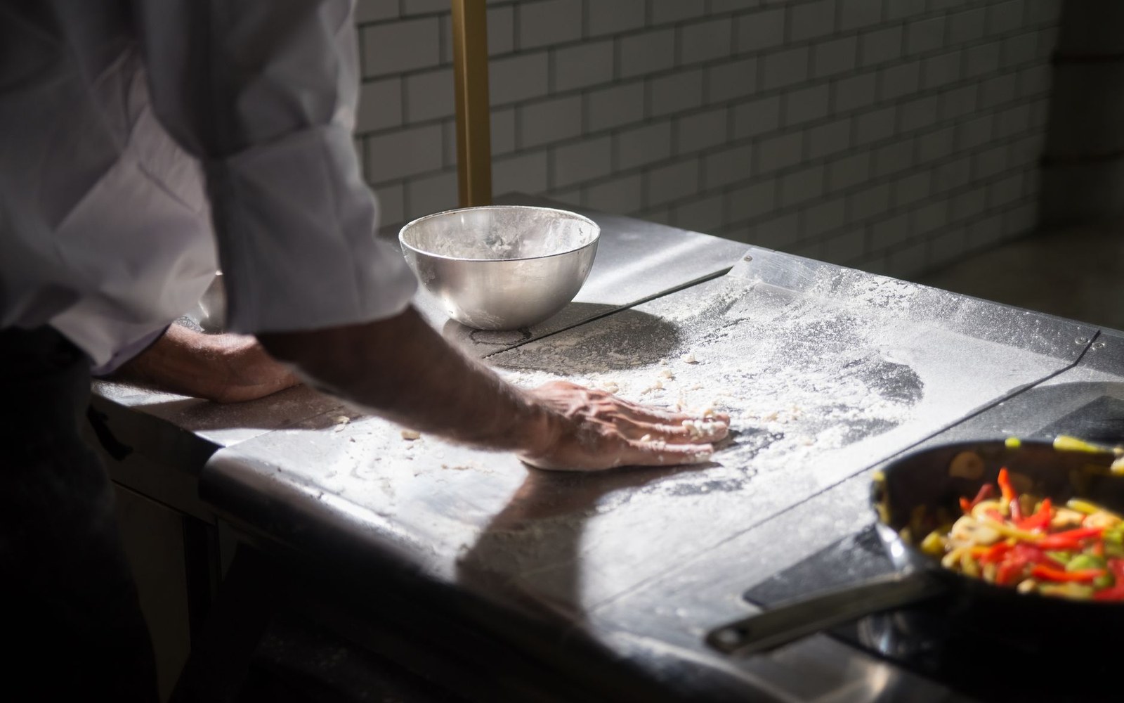 Flour and a metal bench scraper on a stainless steel workbench, a shaped dough piece in the background slightly out of focus
