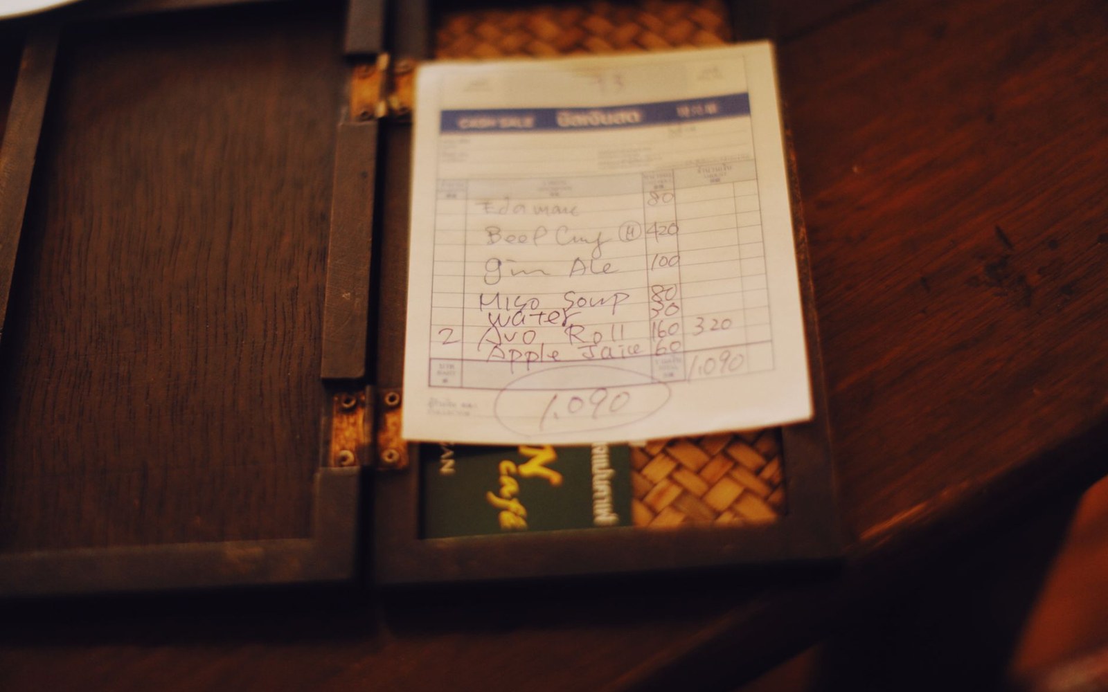 A handwritten restaurant bill listing menu items and prices in a wooden check holder