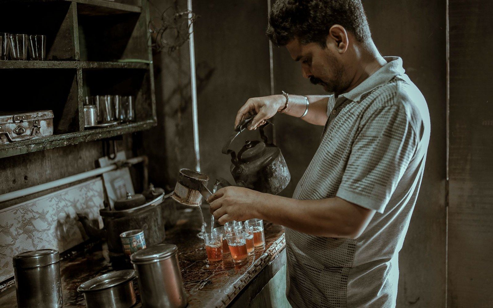 A man pours tea from a metal kettle into small glasses on a worn counter