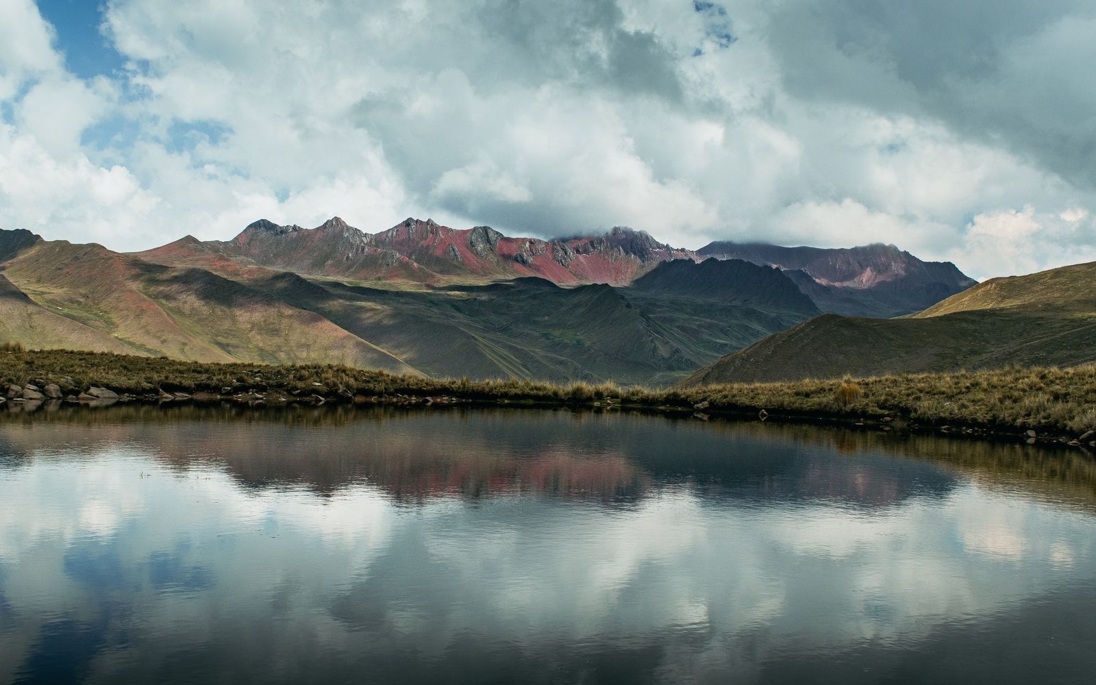 Reddish mountain ridges reflected in a still Andean lake beneath cloudy skies