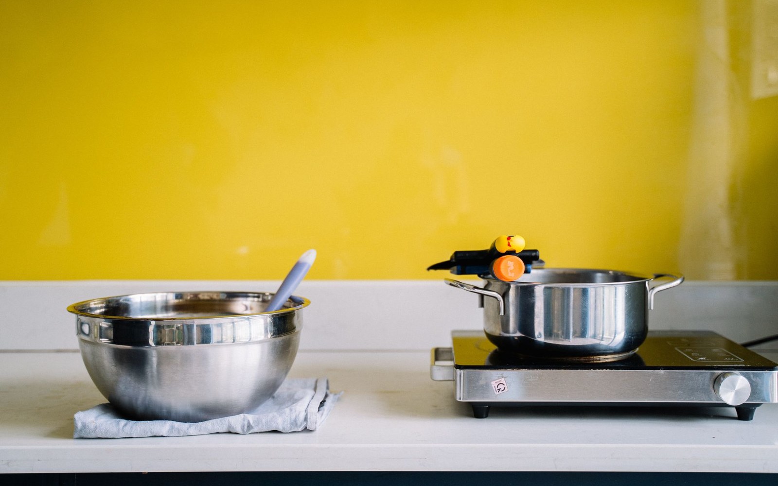 A metal mixing bowl and a small pot on a portable burner against a yellow wall