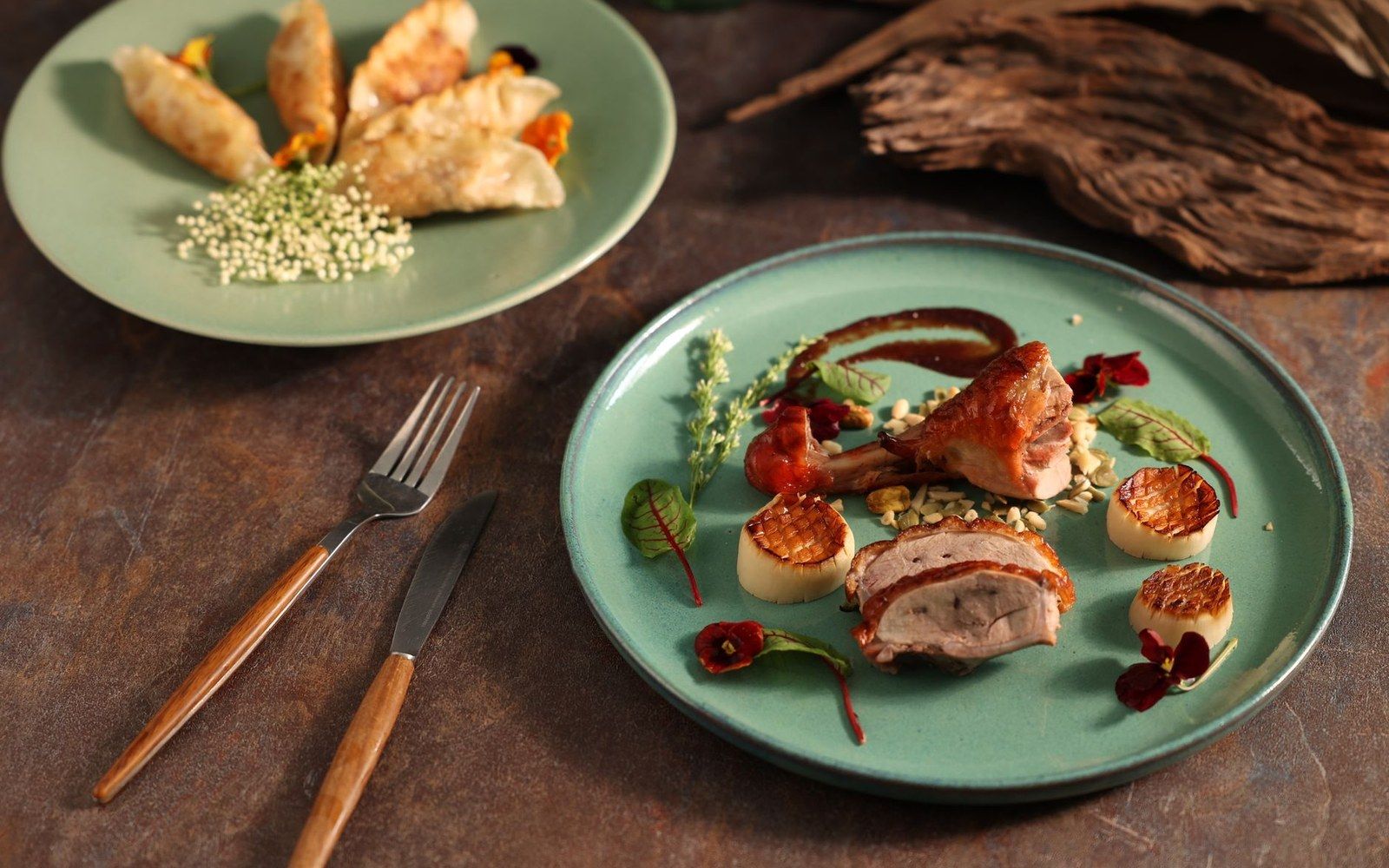 Plated roasted meat with grains, scallops and edible flowers beside a dish of dumplings
