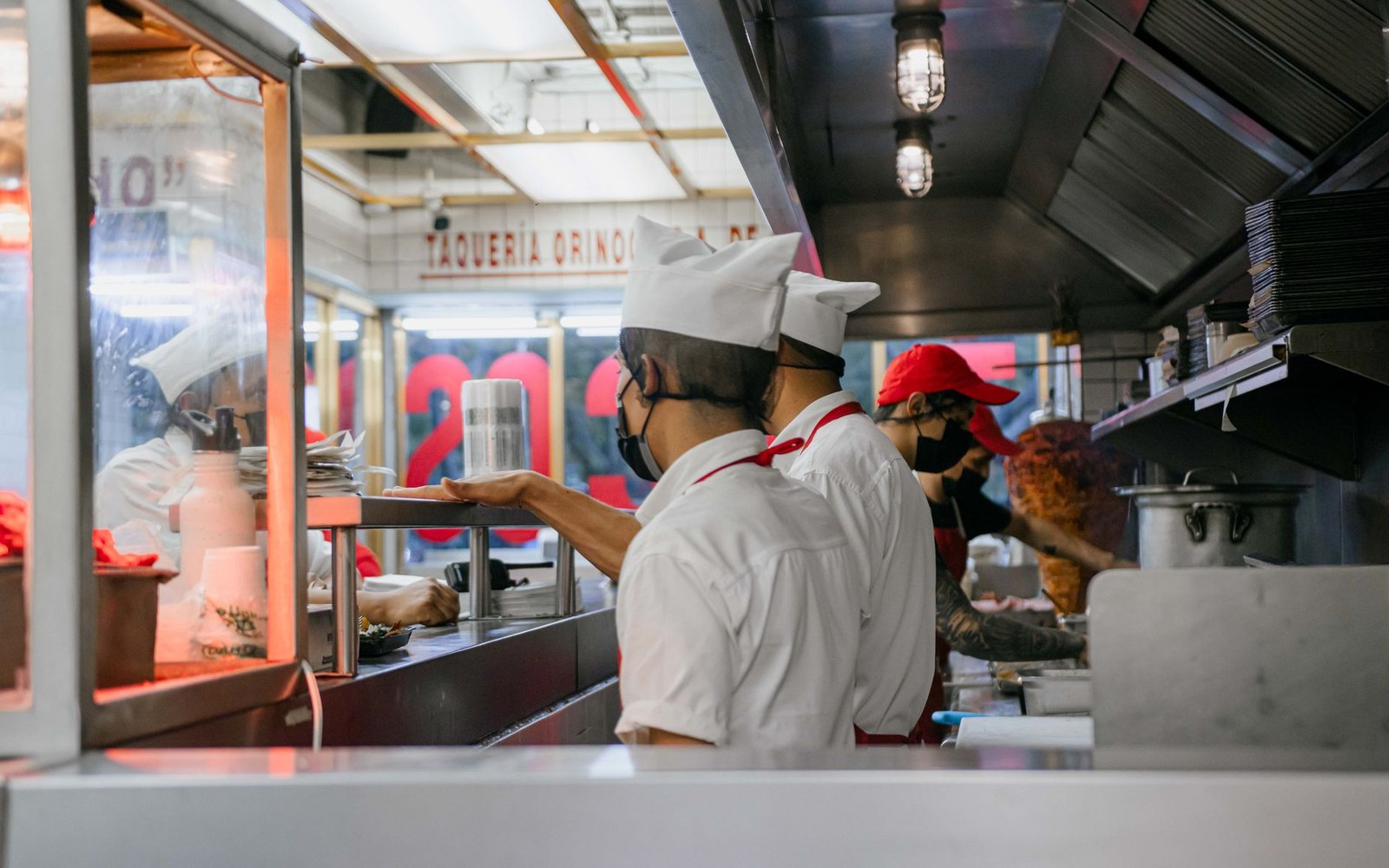 Masked workers prepare tacos beside a spinning trompo at a busy taqueria counter
