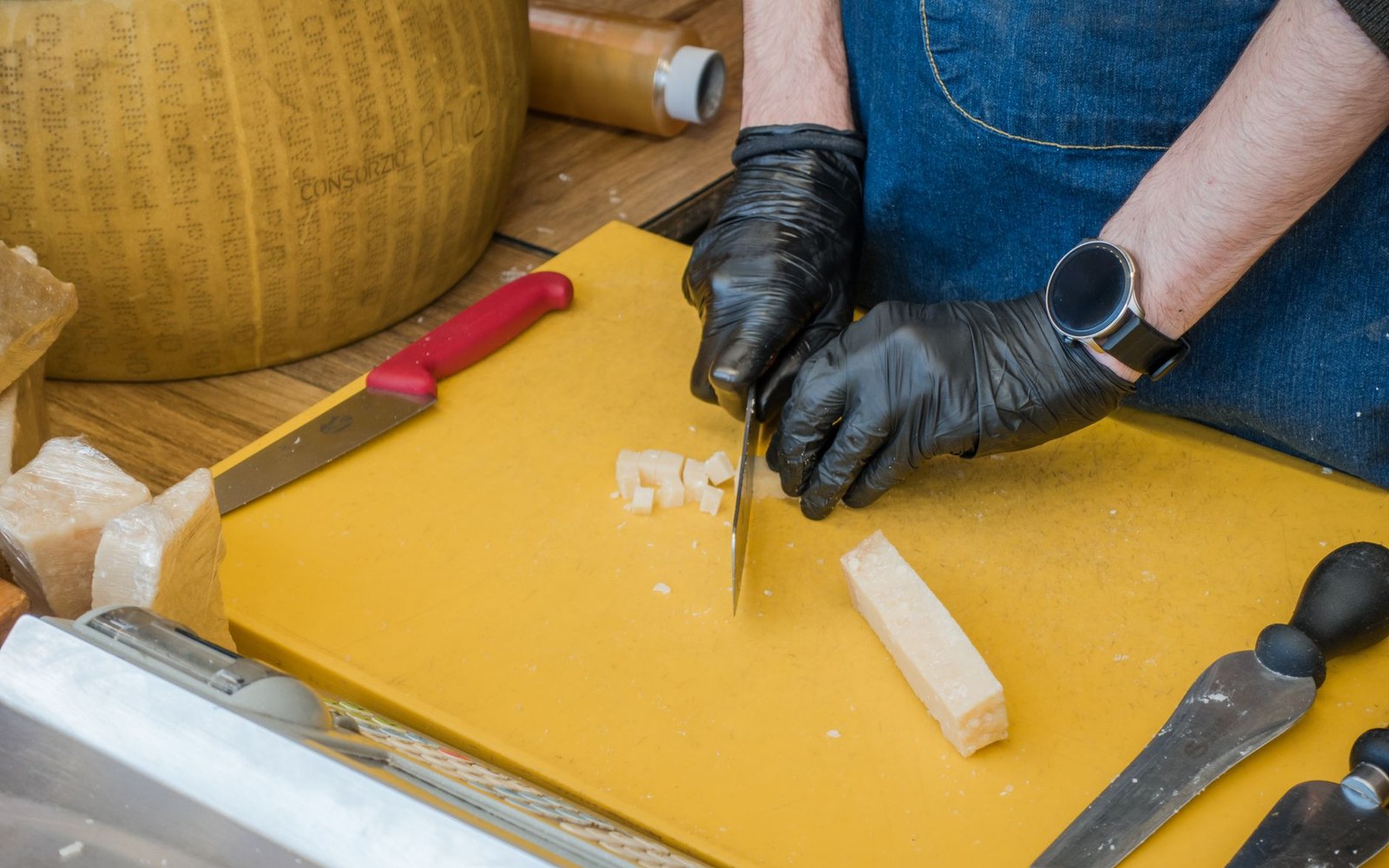 Gloved hands cut a wedge of parmesan cheese into small cubes on a board
