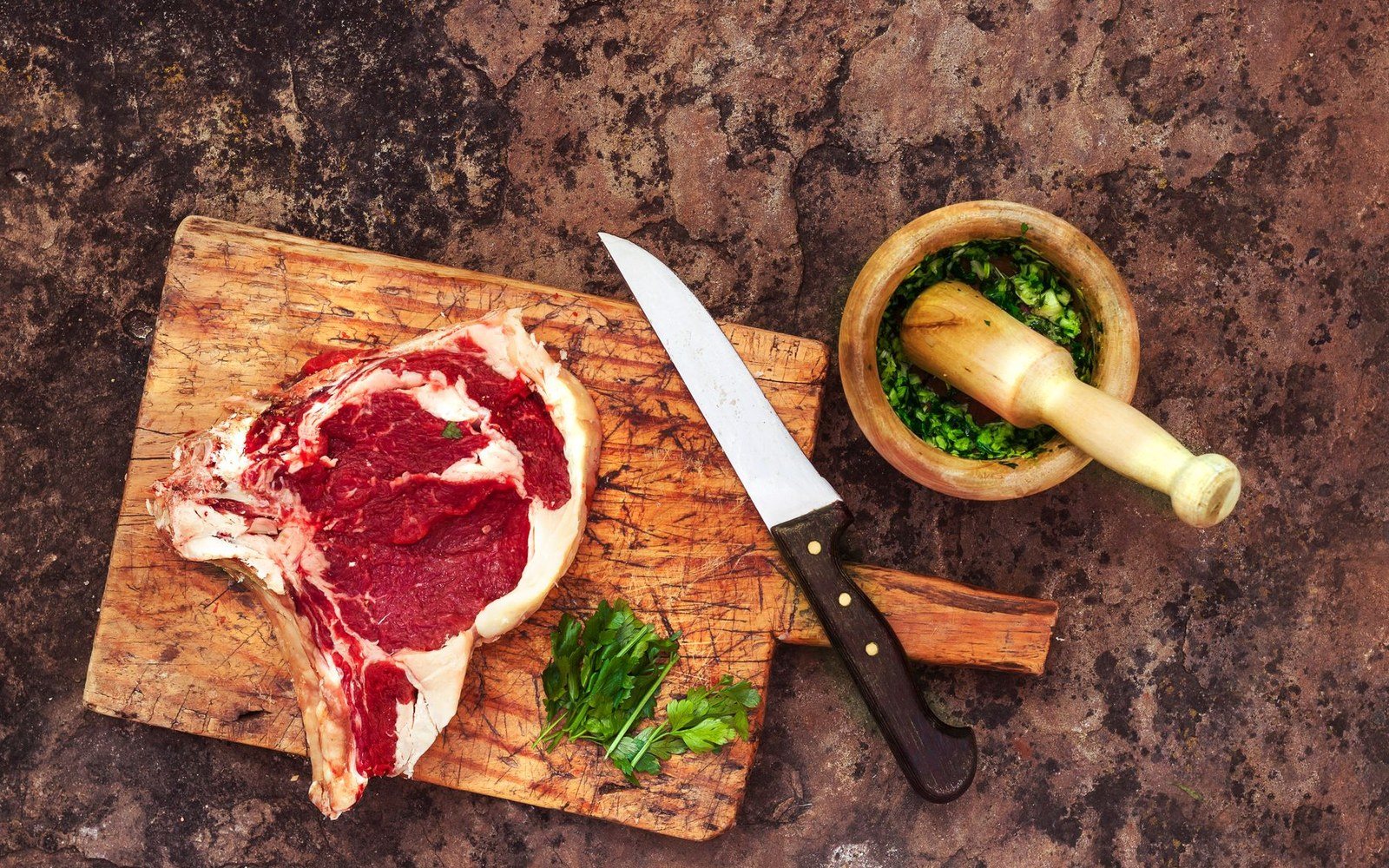 An adult cook's hands working a boning knife along the blade bone of a lamb shoulder on a wooden block, clean cut visible